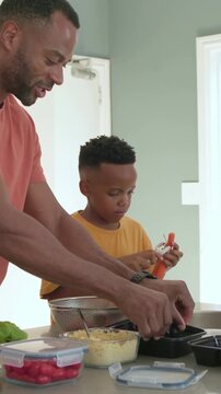 Vertical video: Scooping dad preparing meal trays at kitchen son in mustard tee peeling carrot