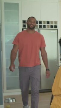 Vertical video: Dad in salmon tee preparing bowls in kitchen, son coming with backpack for hug