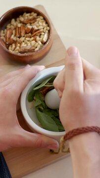 Vertical video: Steadying mortar, hands grinding basil and nuts on kitchen counter for processor