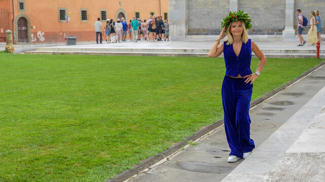 Elegant graduate walking under the Leaning Tower of Pisa wearing a laurel wreath in Piazzale dei Miracoli