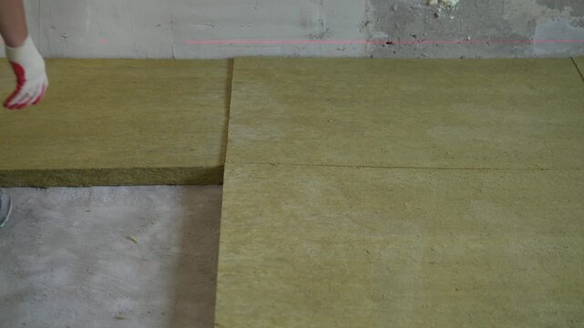 A construction worker carefully lays rigid mineral wool insulation boards on a concrete floor. The process shows floor soundproofing thermal insulation during a home renovation building project