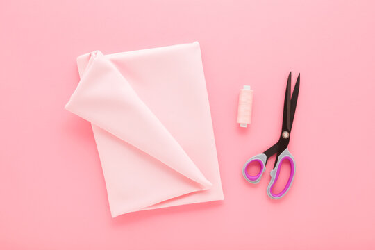 New folded light pink gabardine elastane fabric, thread spool and dressmaker scissors for female clothes sewing on pink table background. Pastel color. Closeup. Top down view.