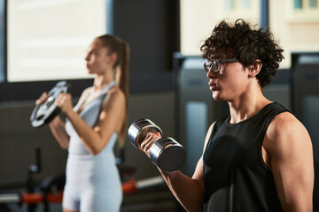 Fototapeta premium Young adult man lifting dumbbell during strength workout while young adult woman training with weight plate in gym. Ideal for fitness marketing, personal training, healthy lifestyle campaigns