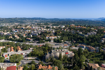 Fototapeta premium Aerial view of Grasse, France, with terracotta roofs, mid rise blocks, cypress and palm trees, winding roads, a curved roundabout, and layered hills under clear skies.
