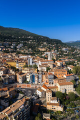 Fototapeta premium A bright aerial view of Grasse, France shows terracotta roofs, a roundabout, and the white bell tower of Grasse Cathedral under clear midday light and deep skies.