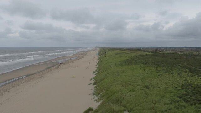 A professional drone shot moves forward over the Dutch coastline, revealing a paraglider entering the frame. The scene captures the grassy dunes and North Sea beach near Zandvoort under a grey sky.