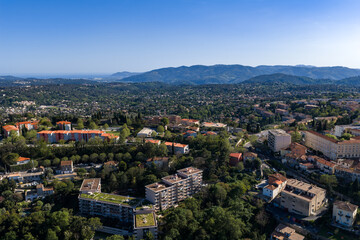 Fototapeta premium Aerial view of Grasse, France shows terracotta roofs, green rooftops, and tree lined streets across a valley, with the Prealps in midday light and rolling hills.