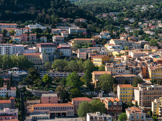 Fototapeta premium Aerial view of Grasse, France shows pastel buildings on green slopes, terracotta roofs, plazas, and narrow lanes, under warm afternoon light with wooded hills.