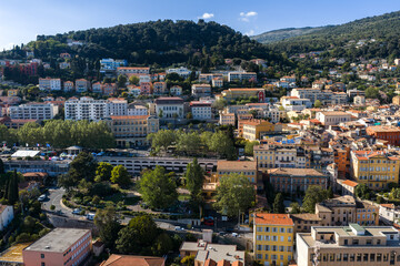 Obraz premium Aerial view of Grasse, France shows pastel buildings on slopes, terraced neighborhoods, narrow streets, and gardens under midday sun, with forested hills and a valley.