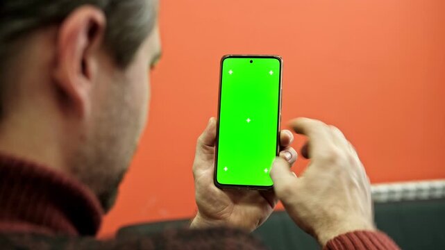 Hand of young man holding smartphone green screen in business office work. Mobile phone vertical mockup. Male person using phone with blank green background display. Chroma key smart phone. Close up.