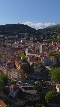 Aerial view of Grasse, France, with cathedral bell tower, pastel roofs, winding streets, plazas, cars and pedestrians, terraced town on a wooded hillside in afternoon light