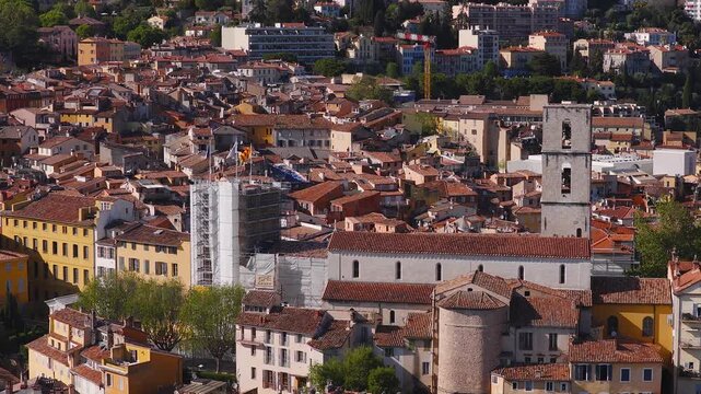 Aerial view of Grasse, Provence, France, as the camera pans across rooftops, a bell tower, a round medieval tower, and a scaffolded site with flags under warm light.