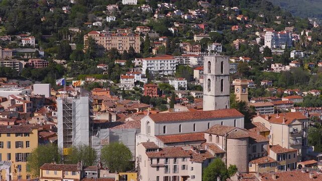 Aerial daytime pan of Grasse, France, shows stone cathedral with square bell tower, terracotta roofs, scaffolded municipal tower with flags, villas and clock tower on hills.