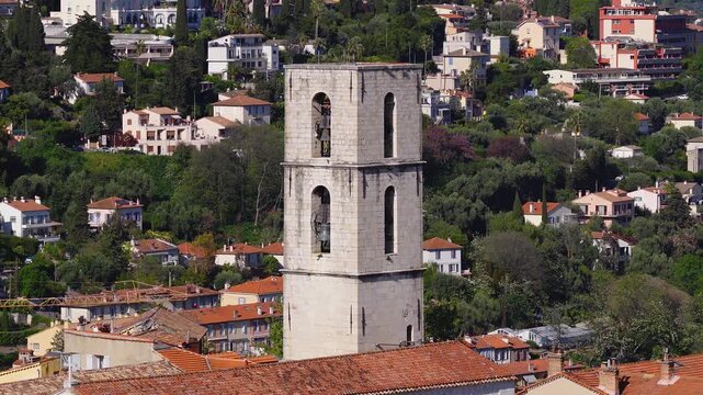 Aerial pan over Grasse, France, centers on a stone bell tower and a pale yellow clock tower above terracotta roofs, with villas, olive trees, and hillside gardens.