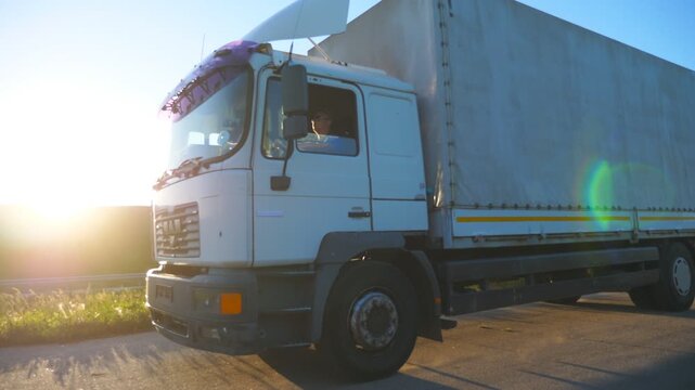 Truck with cargo trailer driving on highway and transporting goods. White lorry riding to destination through countryside at sunset time. Bright sunset light shining at background. Side view Slow mo
