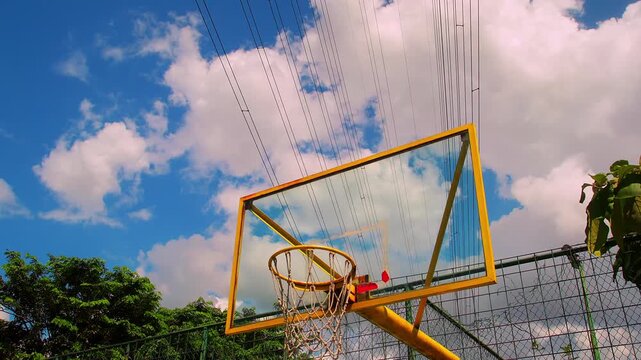 Timelapse of yellow basketball hoop stands tall against blue sky. White clouds drift above the court. Net hangs limp from the rim. The view is bright and open. Ideal for sports, school.
