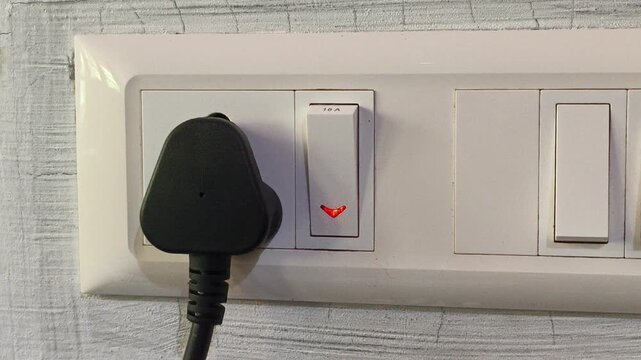 Closeup of finger turn on, pushing or flip plastic electric power switch on indoor wall with red light near socket or outlet with three pin plug. Electricity and energy consumption concept.