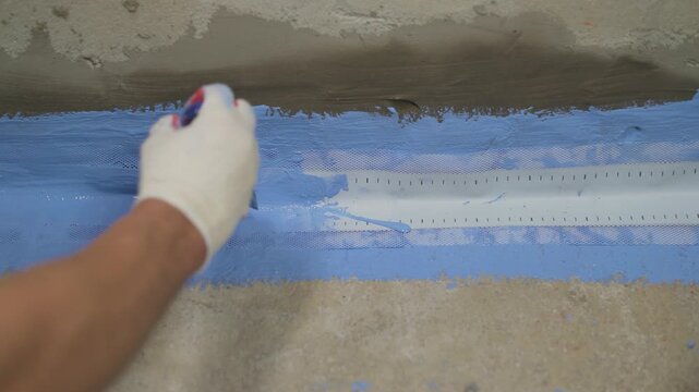 Close up of a construction worker's hand in a glove applying blue liquid waterproofing membrane with a brush over a joint sealing tape on a concrete floor. Home renovation and building process
