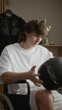 Vertical shot of young Caucasian man in wheelchair putting off moto helmet and taking look at it, remembering old good days