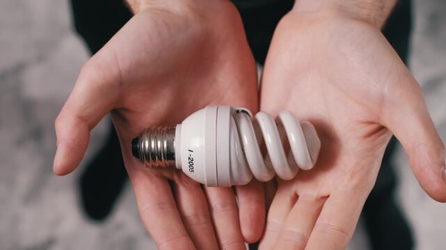 Close-up top view of male hands gently presenting a spiral CFL energy-saving light bulb towards the camera. The zoom emphasizes energy conservation and modern lighting.