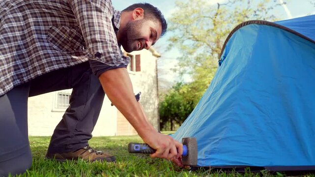Man pitching camping tent hammering stake. Young man assembling a blue tent, using a hammer to drive a stake into the grassy ground