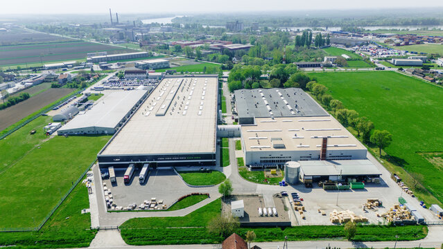 Aerial view of a large industrial warehouse complex with loading docks and trucks surrounded by green fields and trees in Sremska Mitrovica, Vojvodina, Serbia.