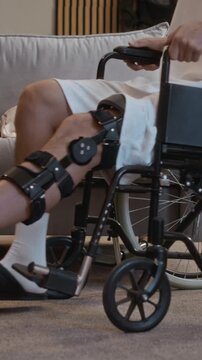 Vertical shot of anonymous young man with injured leg using wheelchair to maintain mobility during recovery at home after moto accident