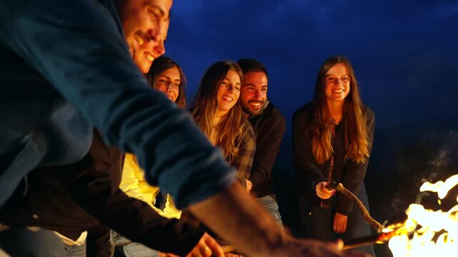 Group of happy friends enjoying a bonfire at night. Young men and women smiling and laughing together around a warm campfire during a cool evening outdoors