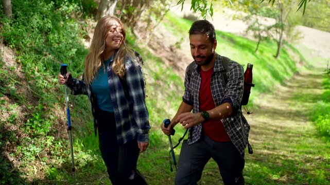 Happy young couple hiking together on a trail in the green forest. Young man and woman hiking with backpacks and walking sticks on a nature trail during sunny day