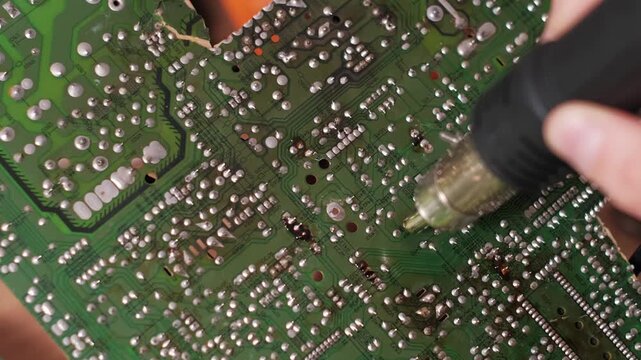 Close-up view zooming out as a technician uses a hot air rework station to heat solder joints on a circuit board. The process demonstrates advanced electronics repair and maintenance.