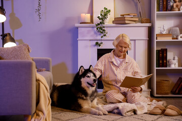 Senior woman with Husky dog reading book near fireplace at home late in evening © Pixel-Shot