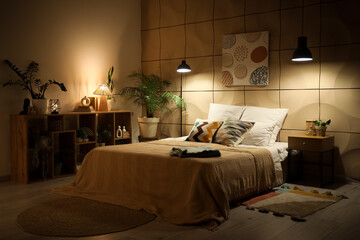 Interior of cozy bedroom with blankets on bed, shelf unit and glowing lamps at night © Pixel-Shot