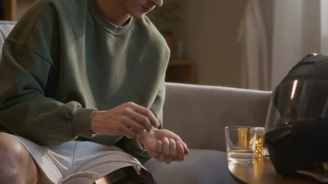 Tilt up shot of young ex motorcyclist drinking water and taking painkillers while resting on sofa in living room at home