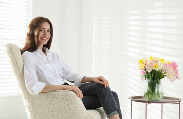Obraz premium Happy woman in armchair near table with beautiful flowers indoors