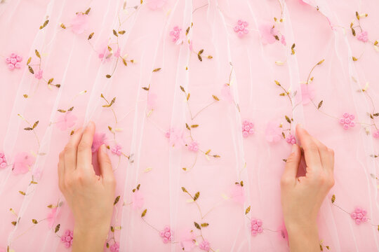 Young adult woman hands touching new transparent soft wrinkled tulle fabric with flower pattern on light pastel pink background. Closeup. Point of view shot. Flat lay. Top down view.