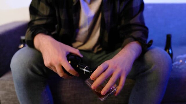 Close-up of a lonely depressed man pouring alcohol from a bottle into a glass while sitting on a sofa. Concept of alcoholism, severe addiction, unemployment, and divorce. High quality 4k footage