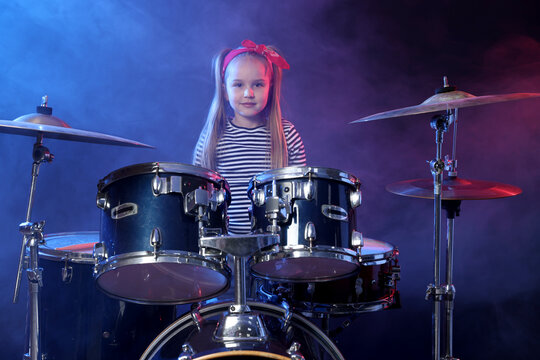Little girl in bandana learning how to play drums in color lights and smoke against dark background