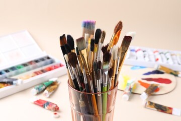 Fototapeta premium Paintbrushes in glass, paints and palette on beige background, selective focus