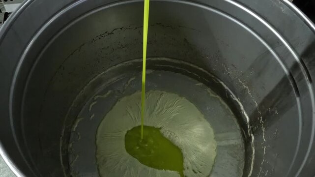 Fresh, cold-pressed olive oil flowing into a large container during the extraction process, showing a golden liquid with natural foam pul, highlighting traditional and industrial olive oil production.