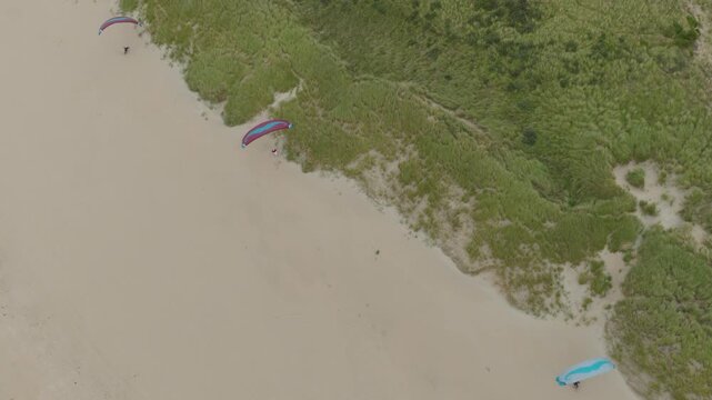 A professional drone shot descends while the camera tilts upward, tracking paragliders moving straight over the dunes. The motion creates a parallax effect against the Dutch coast near Zandvoort.
