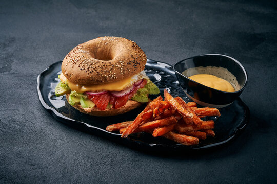 Smoked salmon bagel with avocado, radish, and melted cheese, served with sweet potato fries and dipping sauce on dark plate