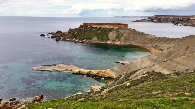 Qarraba Bay in Mġarr Malta with blue clay slopes and limestone headland shaping a small cove over clear Mediterranean water within Ġnejna Bay system, active erosion landscape, forward drone flyover