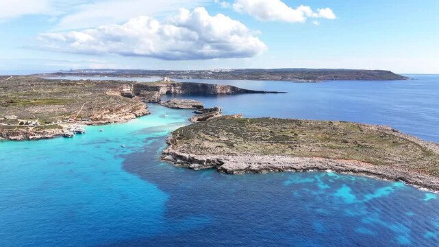 Blue Lagoon in Comino Malta with shallow turquoise water over limestone seabed between Comino and Cominotto islands, clear Mediterranean sea and rugged coast, oblique pullback drone shot
