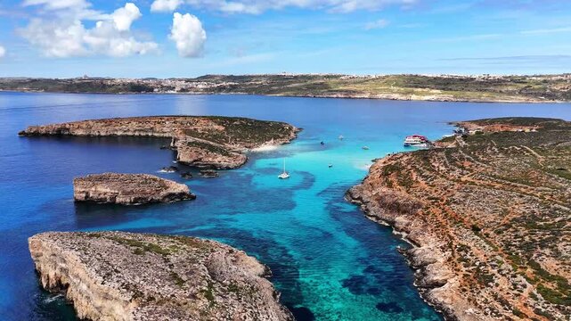 Blue Lagoon Bejn il Kmiemen Comino Għajnsielem Malta showing channel toward Cominotto Kemmunett with shallow limestone seabed and reef patches in Mediterranean Sea, forward panoramic drone shot