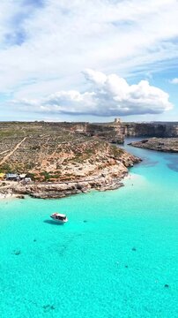 Blue Lagoo Comino Malta with turquoise shallow water, leisure shuttle boat anchored, coastal access path on garigue terrain and St Mary Tower fortification in background, vertical aerial shot