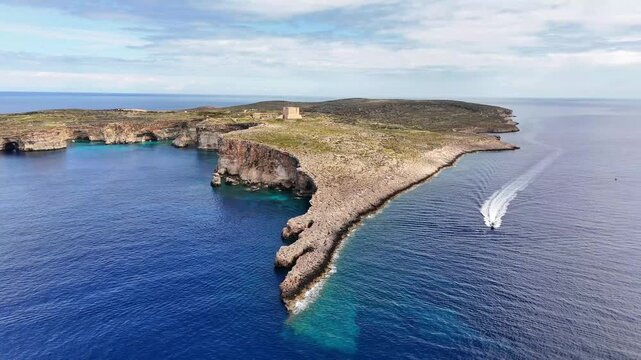 Lantern Point in Comino Malta southwestern tip with Upper Coralline Limestone cliffs and St Mary Tower Torri ta Santa Marija above Mediterranean Sea as motorboat approaches leaving wake, slow forward