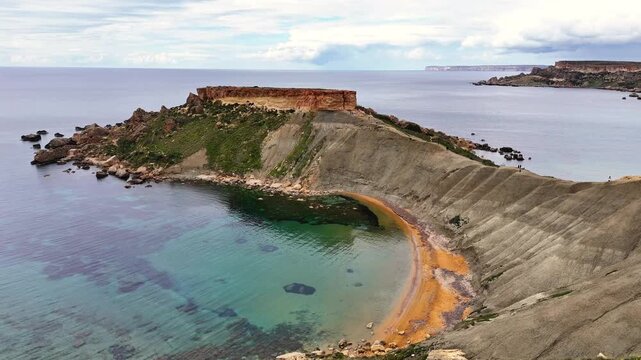Aerial view of rugged coastal cliffs and a curved bay with clear water at Qarraba Bay, Malta, showing rocky shoreline and steep terrain under soft daylight.
