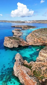 Blue Lagoon in Comino Għajnsielem Gozo Region Malta with coralline limestone cliffs over shallow turquoise basin with Posidonia oceanica habitat and cumulus humilis clouds, forward drone shot flyover