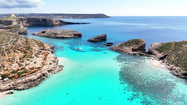 Blue Lagoon Comino Għajnsielem Malta with narrow channel toward Cominotto Kemmunett showing shallow limestone seabed reef patches and anchored sailboat in clear Mediterranean water, drone pullback