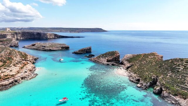 Drone aerial of Blue Lagoon in Comino, Malta showing small boats scattered across bright turquoise water, with rugged rocky shoreline and open sea in daylight.
